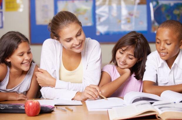 Professora e três alunos sentados à mesa analisam um caderno e sorriem. Professora faz um apontamento no caderno com um lápis.