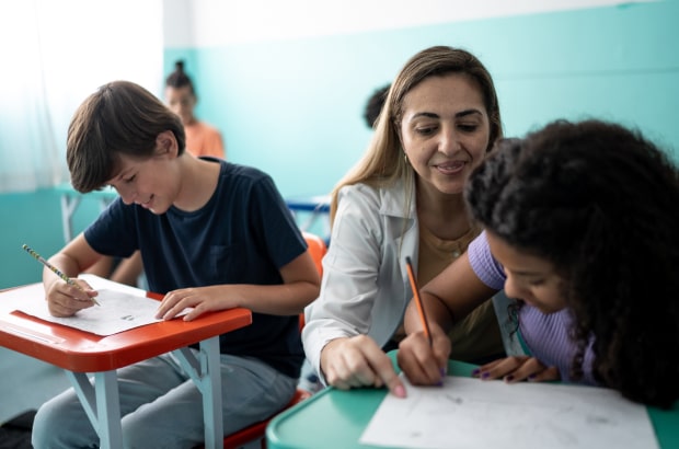 Professora de pé sorri e aponta caderno do aluno, que está sentado, escrevendo.